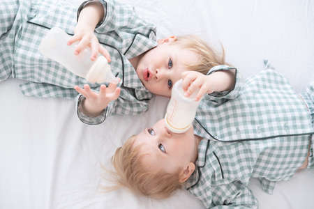 Two toddler baby twin boys in pajamas lie on bed drinking milk from bottles. Candid home childhood...