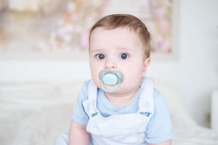 close up portrait baby boy 6 months old with nipple in blu clothes and sitting on white bed at homeの写真素材