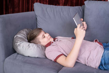 Young teenager boy using tablet lying on gray sofa at home.
