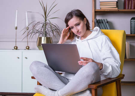 Beautiful young woman in white hoodie using laptop sitting on yellow armchair at home