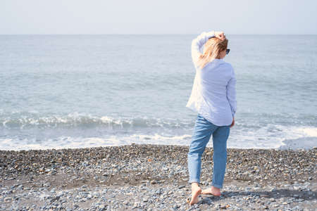 Rear view blond woman standing on shore of sea on sunny day in spring, summer