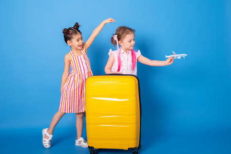 Happy excited little girls friends in colorful clothes with toy plane and yellow suitcase on blue backgroundの写真素材