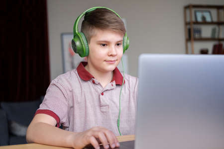 handsome teenager boy in headphones studying online, playing in video games using laptop sitting on...