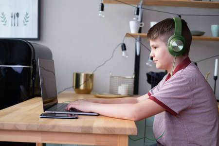 handsome teenager boy in headphones studying online, playing in video games using laptop sitting on...