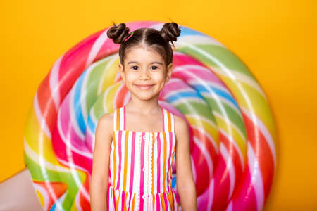 Happy excited arab child girl in colorful striped dress having fun on yellow background with lollipop.の写真素材