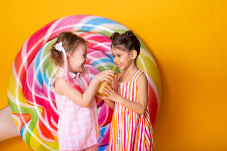 two happy little girls in colorful dress drinking orange juice having fun on yellow background with lollipop.の写真素材