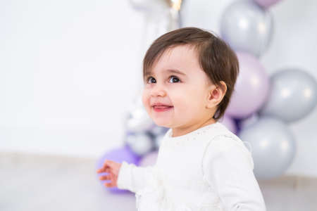 baby girl in white dress celebrating her first birthday with cake and balloons.