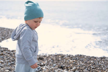 child blond boy playing with rocks and sand on the beach on a sunny day in spring or autumnの写真素材