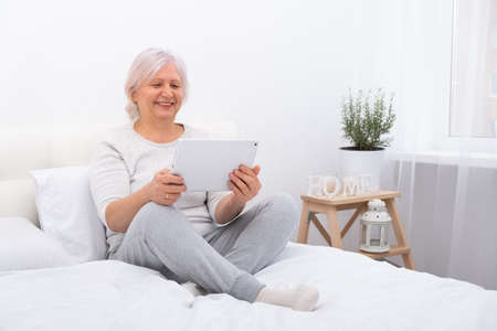 handsome senior women sitting in bed using tablet at home, having video call with children