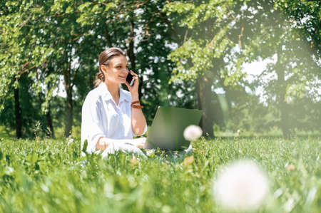 attractive business women in white shirt talking on phone using laptop sitting on grass in park