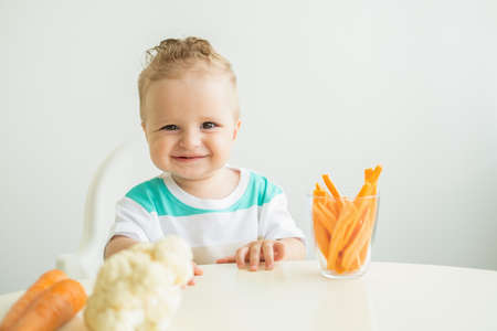 Baby boy sitting in a Childs chair eating carrot slices on white background.の写真素材