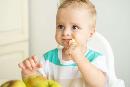 cute baby boy sitting at the table in child chair eating apple on white kitchen.の写真素材