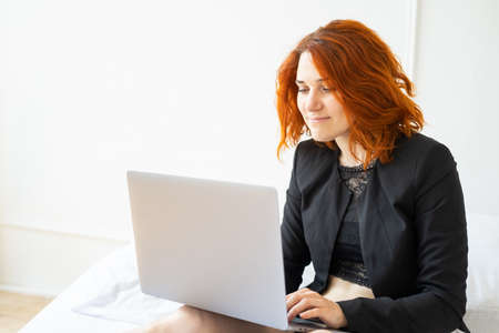beautiful smiling ginger woman middle age sitting on bed using laptop in light roomの写真素材