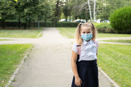 blonde curly schoolgirl in protective mask in school uniform with pink backpack back to school.の写真素材
