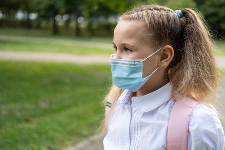 portrait of blonde curly schoolgirl in protective mask in school uniform with pink backpack back to...