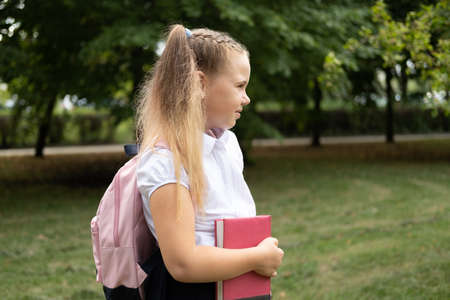 blonde smiling schoolgirl in school uniform holding notebook with pink backpack back to school outdoor.の写真素材