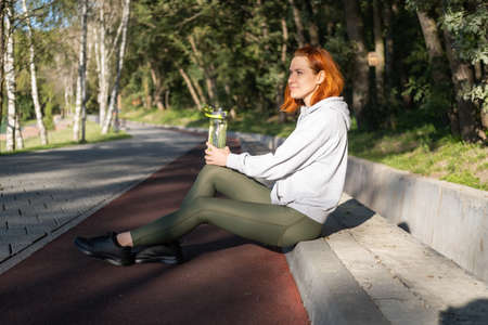 slim ginger woman runner in sport clothes sitting in park, drinking water. Healthy fitness lifestyleの写真素材