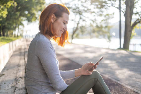 slim ginger woman in sport clothes using mobile phone at park. Health, wellness, fitness lifestyleの写真素材