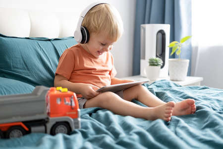 Smiling happy little kid boy using tablet in wireless headphones in bed at home.の写真素材