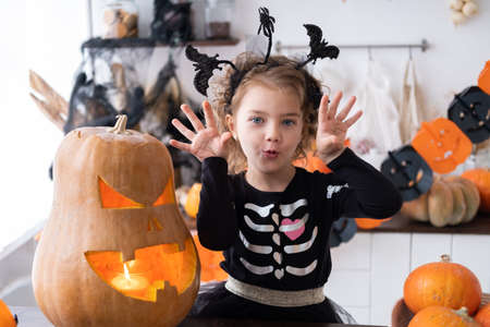cute girl in costume of witch with pumpkin at home in kitchen, having fun, celebrating Halloween.の写真素材