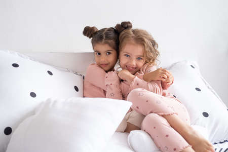 two beautiful diverse kids girls in pajamas hugging on bed in modern bright apartment.の写真素材