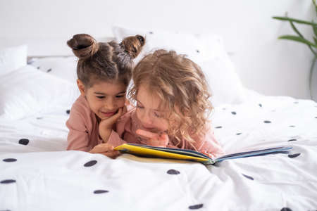 Two diverse kids girl sisters friends in pajamas reading book lying on white bedding on bed at home.の写真素材