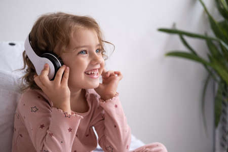 Smiling happy little kid girl in pajama with wireless headphones listening music in bed at home.