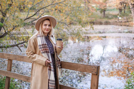 attractive young woman in beige coat and hat drinks coffee by the lake in autumn park in fallの写真素材