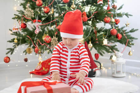Baby girl in striped bodysuit and Santa hat playing with gift box near Christmas tree