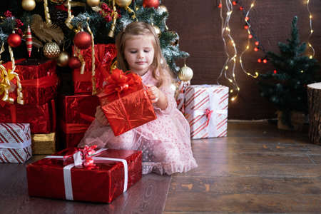 Happy little smiling blond girl in beautiful dress with gift box near christmas treeの写真素材