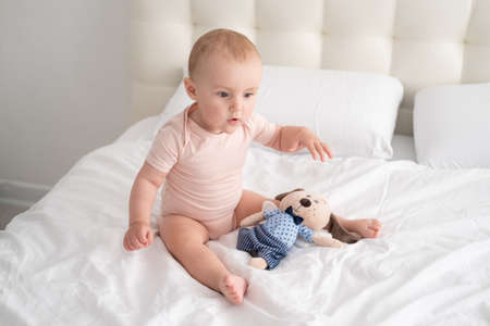 baby girl in light pink bodysuit playing with soft toy on white bedding on bedの写真素材