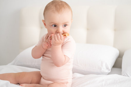 baby girl in light pink bodysuit nibbling wooden toys on white bedding on bedの写真素材