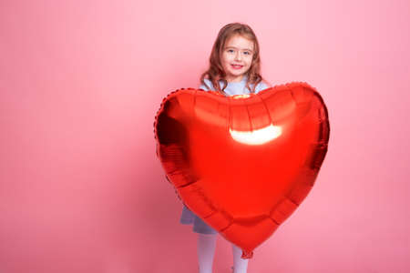 Beautiful child girl with red heart balloon on pink background celebrating Valentine dayの写真素材