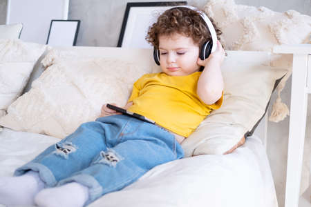 Smiling happy kid curly girl using phone in wireless headphones in bed at home