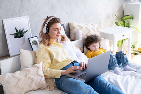 Mother freelancer using laptop in wireless headphones lying on bed with kid daughter using phoneの写真素材