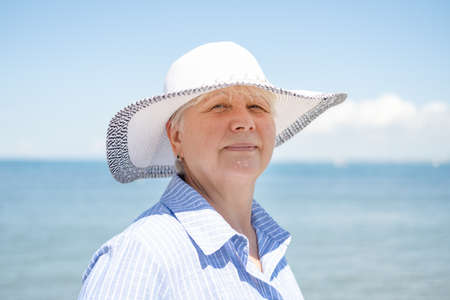portrait of happy senior woman in straw hat and blue shirt staying on seasideの写真素材