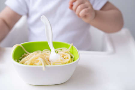 close up cute baby girl eating spaghetti pasta sitting in baby chair on white kitchenの写真素材