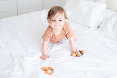 baby girl with blue eyes in brown bodysuit playing with wooden toys on white bedding on bed