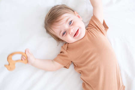 baby girl with blue eyes in brown bodysuit playing with wooden toys on white bedding on bed