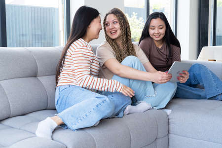 Happy young carefree diversity women friends in casual using tablet, having fun together at home