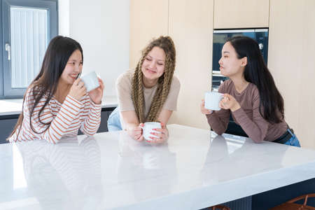 three happy young diversity girlfriends drinking tea, talking, having fun together at modern kitchenの写真素材