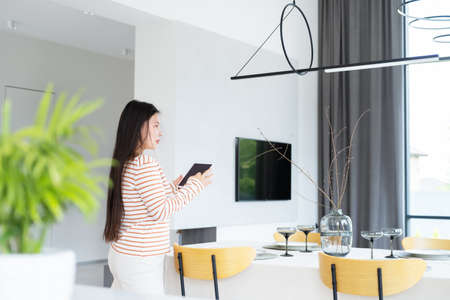 asian woman controlling smart home functions through tablet at modern home, looking at ceiling lampの写真素材