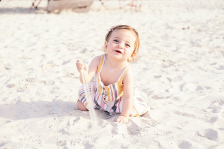 happy baby girl sitting on the beach on sand wearing striped summer dress on sunny day
