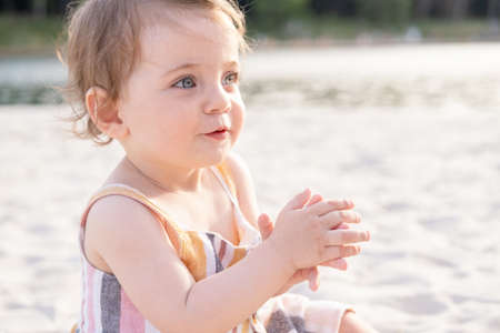 happy baby girl sitting on the beach on sand wearing striped summer dress on sunny day