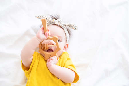 healthy asian baby girl in yellow bodysuit plays with wooden toy teether for teeth on white beddingの写真素材