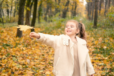 smiling happy little child girl in coat and dress holding autumn leaves, having fun in fall forestの写真素材