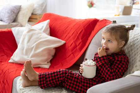 little girl in red pajama holding cup of cacao with marshmallows sitting on couch in living roomの写真素材