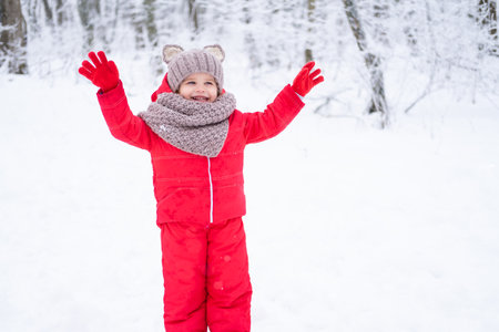 Cute little girl in pink snowsuit and knitted hat and scarf plays with snow in winter forest.の写真素材