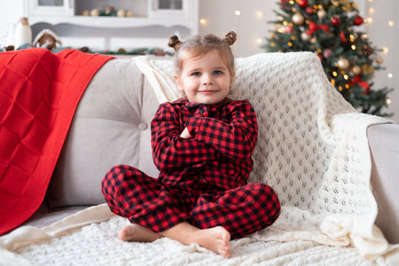 cute little girl in red Christmas pajama smiling sitting on couch.の写真素材
