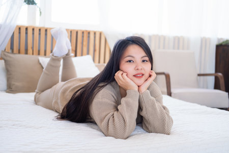 young smiling beautiful gen z asian women with long brunette hair in casual clothing lying on bed at home.の写真素材